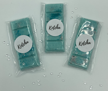 Load image into Gallery viewer, The picture is of a wax melt in the shape of a chocolate bar. The colour is green and blue (teal) with silver biodegradable glitter wrapped in plastic with the brand name "Kotcha" on it. There are crystals surrounding the image and a white background.
